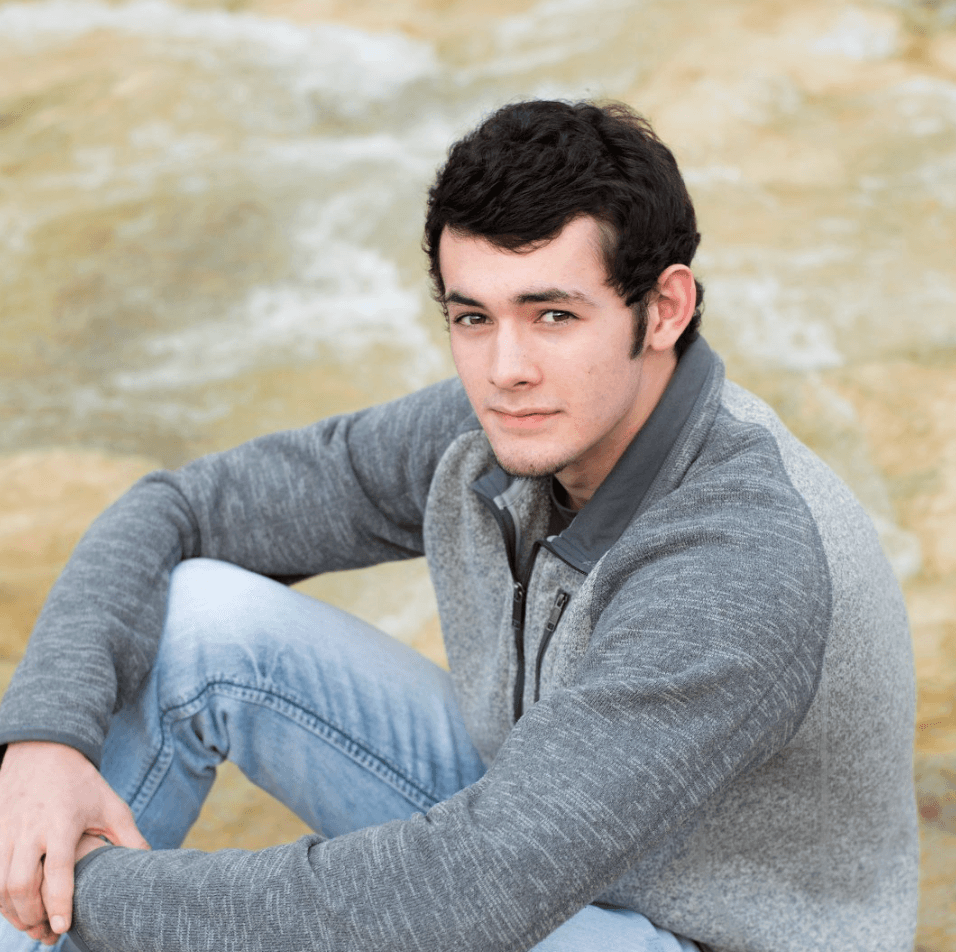 Young man with dark curly hair wearing a grey zip-up sweater sitting outdoors.